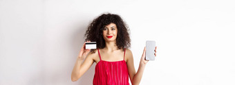 在线购物时尚女人卷曲的头发穿红色的衣服显示智能<strong>手机屏</strong>幕塑料信贷卡微笑相机白色背景