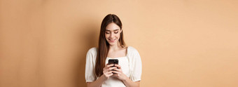 年轻的女人发短信<strong>消息</strong>移动电话微笑阅读智能手机屏幕站米色背景