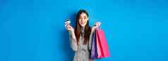 兴奋美丽的女模型购物塑料信贷卡持有商店袋微笑快乐相机蓝色的背景