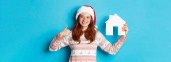 假期<strong>广告片</strong>真正的房地产概念满意女模型红色的波浪头发穿圣诞老人他毛衣显示纸房子模型竖起大拇指蓝色的背景