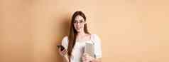 微笑年轻的女人眼镜阅读令人兴奋的新闻移动电话持有移动PC快乐相机站米色背景