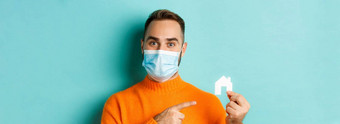 真正的房地产冠状病毒流感大流行概念特写镜头成人男人。医疗面具指出小纸房子站光蓝色的背景