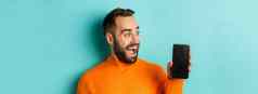 特写镜头年轻的有胡子的男人。显示电话屏幕惊讶穿橙色毛衣站工作室背景