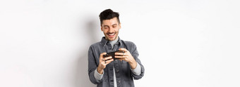 英俊的年轻的男人。玩移动视频游戏微笑屏幕站白色背景