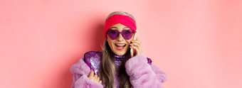 时尚特写镜头时尚的亚洲高级女人听到令人惊异的新闻移动电话微笑快乐相机穿时尚的太阳镜粉红色的背景