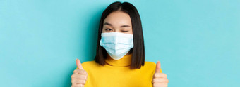 科维德社会距离流感大流行概念可爱的亚洲女孩医疗面具眨眼相机显示拇指好工作手势赞美不错的工作蓝色的背景