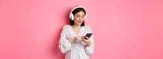 美丽的亚洲女人发短信消息智能手机听音乐耳机站粉红色的背景