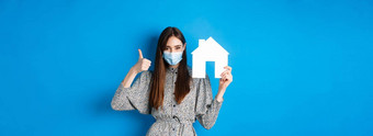 真正的房地产科维德流感大流行概念坦诚的女人医疗面具显示拇指纸房子断路配给公寓站蓝色的背景