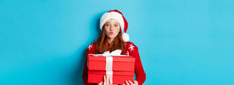快乐假期圣诞节概念可爱的红色头发的人女孩持有礼物皱纹嘴唇吻穿圣诞老人他有趣的毛衣蓝色的背景