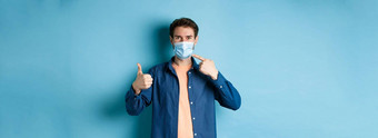 冠状病毒流感大流行概念年轻的健康的男人。指出医疗面具显示拇指预防措施捕捉科维德蓝色的背景