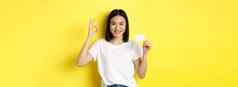 年轻的亚洲女人休闲白色t恤显示塑料信贷卡手势推荐银行微笑相机黄色的背景