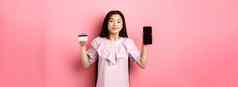 快乐的亚洲女孩显示空智能手机屏幕信贷卡购物在线站粉红色的背景