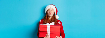 快乐假期圣诞节概念可爱的红色头发的人女孩持有礼物皱<strong>纹</strong>嘴<strong>唇</strong>吻穿圣诞老人他有趣的毛衣蓝色的背景