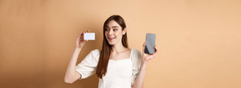 年轻的高加索人女人显示塑料信贷卡很高兴微笑演示空手机屏幕站米色背景