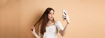 时尚的年轻的女人玩头发采取自拍智能手机使<strong>照片社会</strong>媒体站米色背景