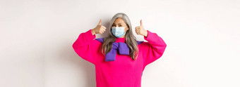 科维德流感大流行社会距离概念快乐的时尚的亚洲高级女人穿医疗面具显示竖起大拇指站白色背景