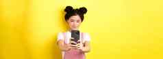 时尚的亚洲女孩采取照片智能手机使视频手机微笑站黄色的背景