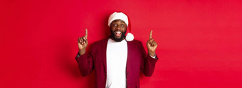 圣诞节聚会，派对假期概念兴奋黑色的男人。圣诞老人他夹克指出手指微笑显示促销提供红色的背景