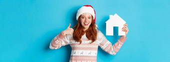 假期广告片真正的房地产概念满意女模型红色的波浪头发穿圣诞老人他毛衣显示纸房子模型竖起大拇指蓝色的背景