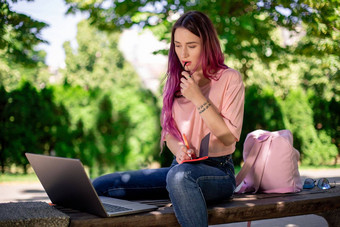 女人写作笔记本坐着木板凳上公园女孩工作在户外可移植的电脑复制空间
