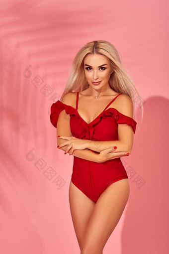 惊人的金发女郎女模型令人惊异的身体站优雅的红色的泳衣粉红色的背景棕榈影子