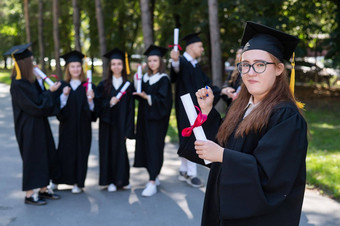 肖像年轻的高加索人女人眼镜研究生礼服背景同学们集团研究生学生在户外