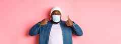 冠状病毒生活方式全球流感大流行概念年轻的非裔美国人男人。指出脸面具显示竖起大拇指保护科维德粉红色的背景