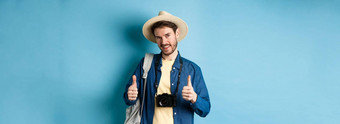 英俊的高加索人旅游稻草他持有背包照片相机显示拇指推荐旅行机构酒店度假胜地蓝色的背景