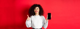 肖像有吸引力的微笑女人卷曲的头发显示空移动电话屏幕翘拇指推荐在线促销站红色的背景
