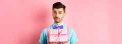 假期庆祝活动概念有吸引力的年轻的男人。胡子穿节日装蝴蝶结显示可爱的礼物盒子相机站粉红色的背景