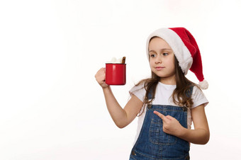 孤立的肖像白色可爱的婴儿女孩圣诞老人他指出红色的杯热可可喝棉花糖