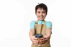 可爱的微笑十几岁的男孩米色老板围裙将生态纸板杯子香外卖咖啡相机