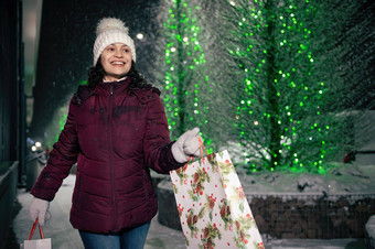 令人愉快的拉美裔女人走购物袋城市街照亮花环雪冬天晚上