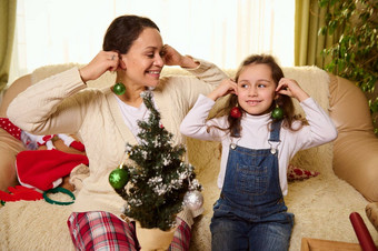 快乐的妈妈。女儿有趣的把闪亮的玩具球耳环装修圣诞节树舒适的首页