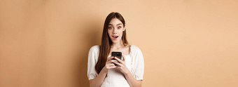令人兴奋的新闻电话惊讶年轻的女人哇阅读在线公告智能手机站米色背景