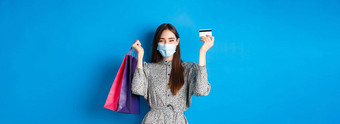 科维德流感大流行生活方式概念漂亮的女模型医疗面具欢乐购物折扣持有塑料信贷卡袋站快乐蓝色的背景