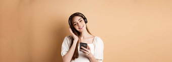 可爱的微笑女孩听音乐无线耳机智能手机站米色背景