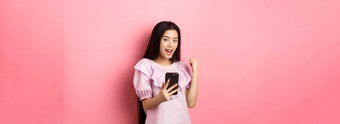 在线购物满意亚洲女孩赢得移动电话使拳头泵持有智能手机站粉红色的背景