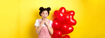惊讶朝鲜文女孩喘气哇特殊的促销提供情人节一天站大红色的心气球黄色的背景