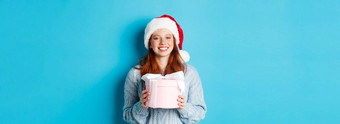 冬天假期圣诞节夏娃概念微笑红色头发的人女孩毛衣圣诞老人他持有一年礼物相机站蓝色的背景