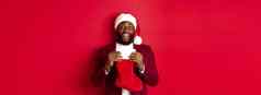 兴奋黑色的男人。开放圣诞节袜子礼物糖果微笑快乐站圣诞老人他红色的背景