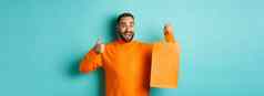 快乐男人。持有橙色购物袋欢乐折扣庆祝显示拇指推荐站蓝色的背景