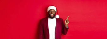 圣诞节聚会，派对假期概念兴奋逗乐黑色的男人。指出手指左微笑显示标志站红色的背景