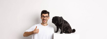 图像快乐满意年轻的男人。坐着哈巴狗狗显示竖起大拇指微笑赞扬好产品白色背景