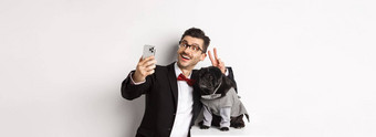 快乐的狗老板西装庆祝圣诞节狗采取自拍智能手机可爱的黑色的哈巴狗服装白色背景