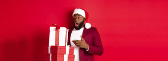 圣诞节一年购物概念惊讶黑色的男人。圣诞节礼物惊讶的是穿圣诞老人他站礼物红色的背景