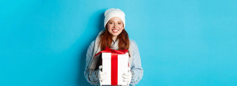 冬天假期圣诞节销售概念可爱的红色头发的人茶水女孩无檐小便帽sewater围巾持<strong>有礼</strong>物包装盒子微笑祝快乐圣诞节站蓝色的背景