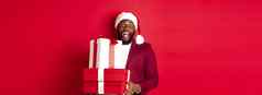 圣诞节一年购物概念快乐的黑色的男人。秘密圣诞老人持有圣诞节礼物微笑兴奋带礼物站红色的背景