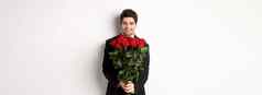 图像英俊的的男朋友黑色的西装持有花束红色的玫瑰微笑日期站白色背景