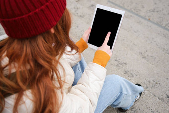 后视图红色头发的人女孩触摸数字平板电脑屏幕触控板文本消息互联网应用程序小工具坐在楼梯在户外
